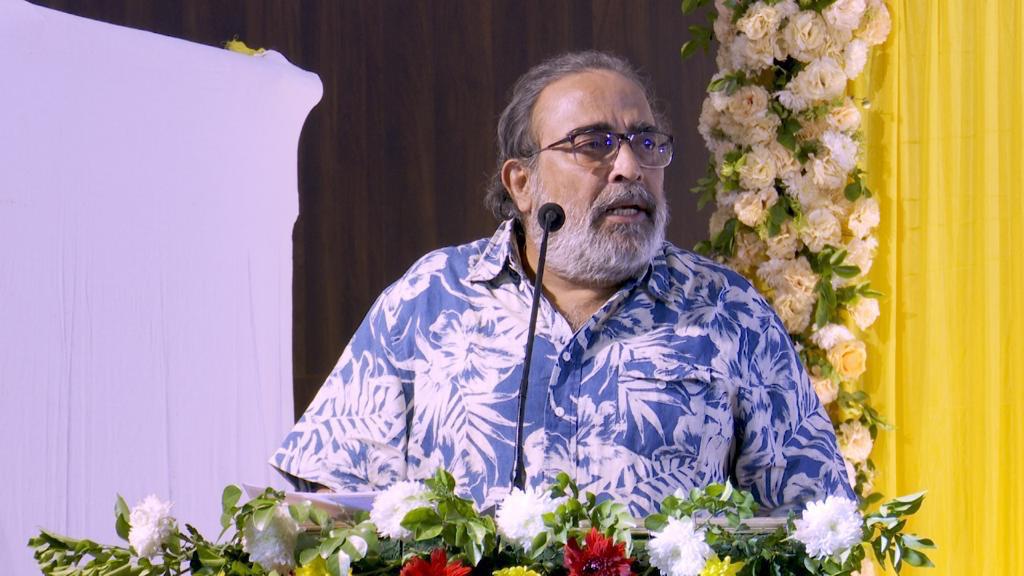 ফুলে সাজানো মঞ্চে মাইক্রোফোনের সামনে চশমা ও নীল ছাপা জামা পরা দাড়িওয়ালা এক ব্যক্তি।